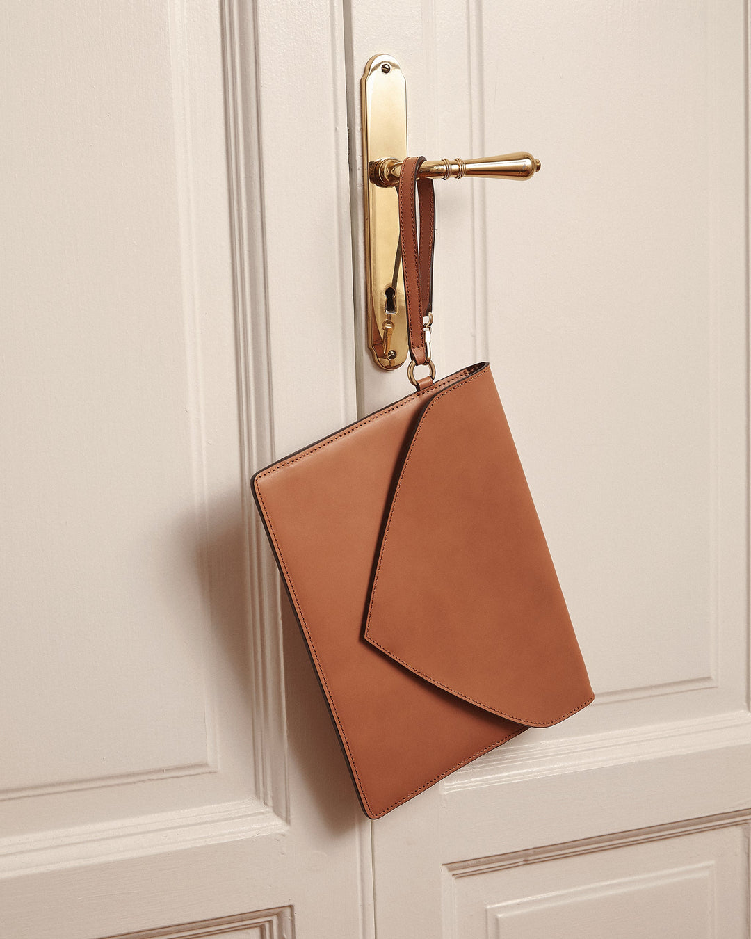 Brown leather clutch bag hanging on a gold door handle against a white paneled door.
