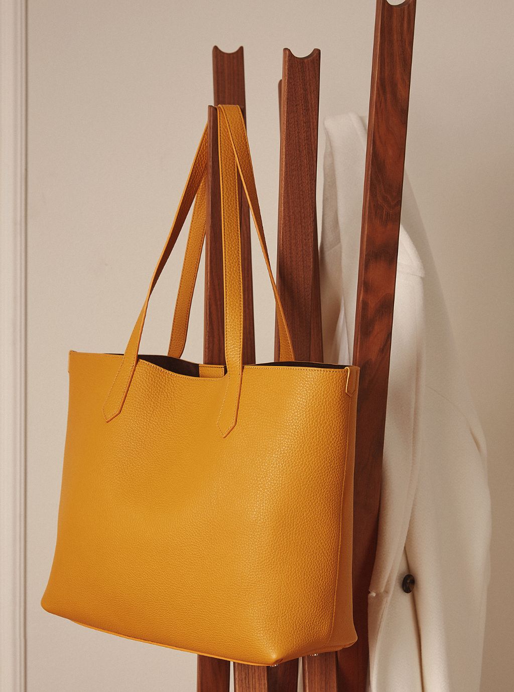A mustard yellow pebbled leather tote bag hanging from a wooden coat rack next to a white coat.