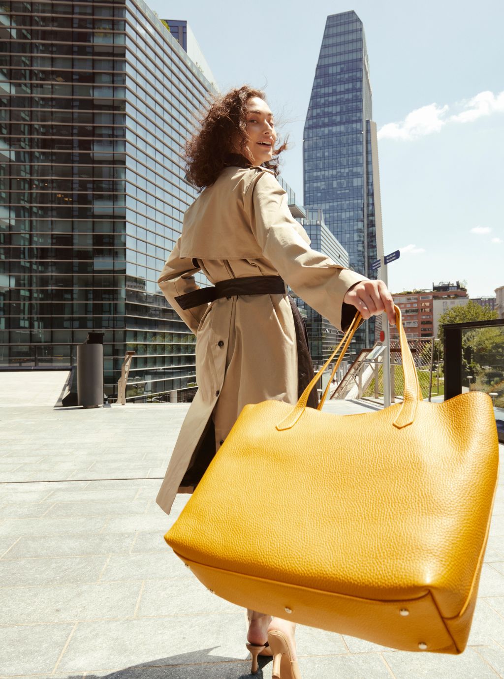 A woman in a beige trench coat carrying a large textured mustard yellow leather tote bag, set against a backdrop of modern glass skyscrapers.