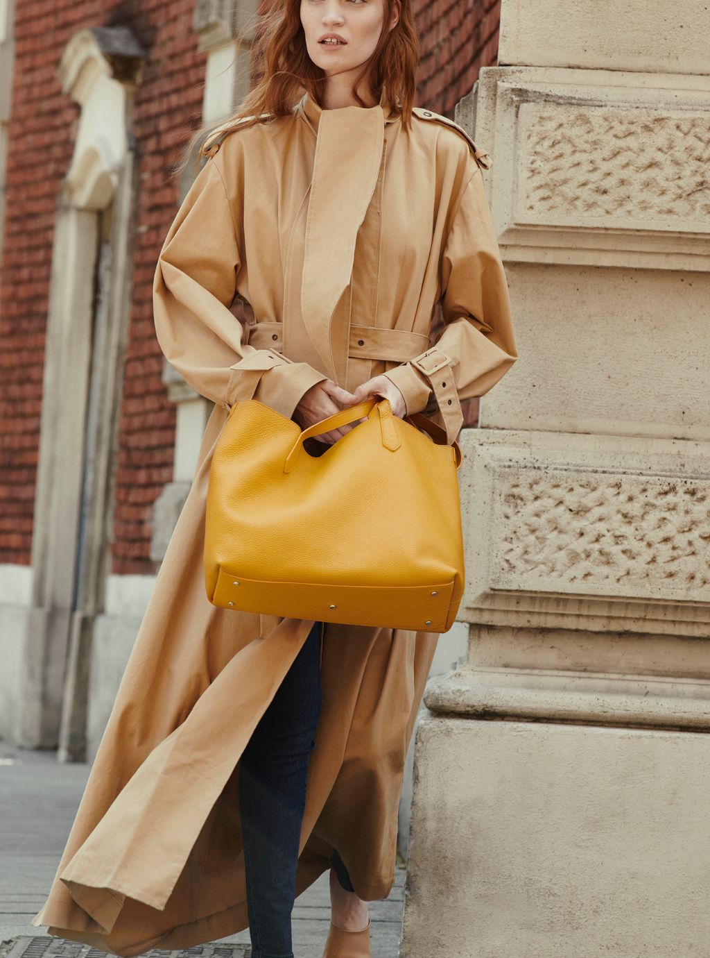 A woman wearing a long tan trench coat and dark blue jeans carries a large mustard yellow leather tote bag while standing outdoors next to a stone column.